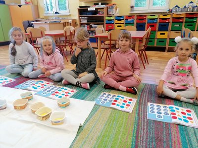Dzieci siedzą na dywanie. Przed nimi znajdują się kolorowe plansze